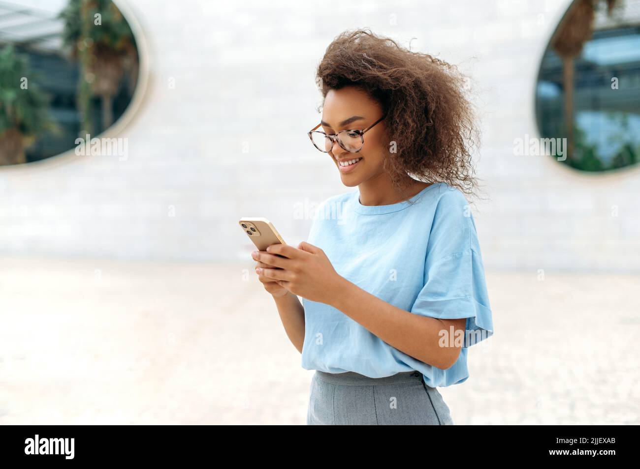 Gadget, tecnologia wireless. Giovane donna riccio di successo afroamericana con occhiali, maglietta blu, in piedi all'aperto vicino al centro affari, usando il telefono delle cellule, inviare SMS, rispondere alle e-mail, sorrisi Foto Stock