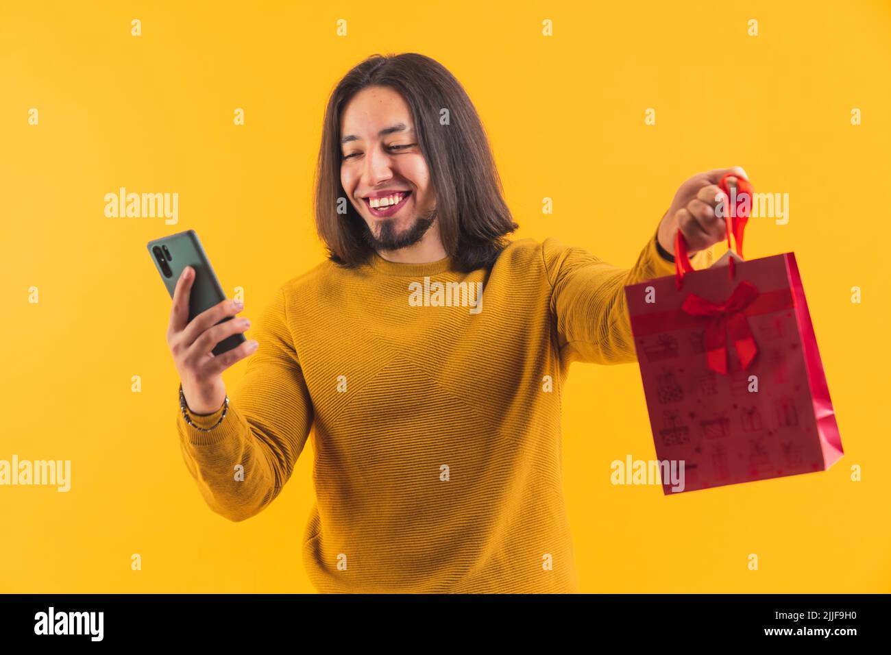 Un lungo pelato bearded giovane uomo felice appeso un telefono e un sacchetto rosso del regalo nella sua mano destra all'interno di una stanza. Foto di alta qualità Foto Stock