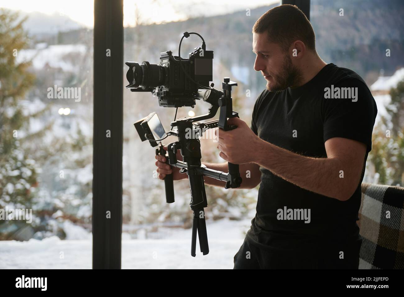 Uomo videografo che realizza riprese, utilizzando una fotocamera montata su un'attrezzatura stabilizzatore gimbal. Finestre panoramiche con paesaggio invernale sullo sfondo. Foto Stock