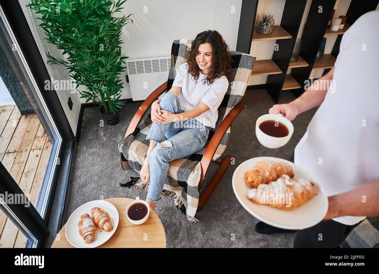 Donna felice seduta in sedia e sorridente mentre l'uomo tiene una tazza di caffè e croissant. Coppia in amore trascorrere il tempo libero insieme gustando la colazione in casa accogliente. Foto Stock