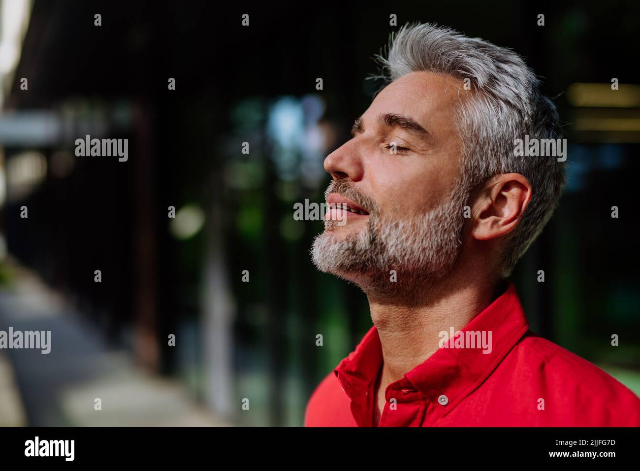 Un uomo d'affari allegro e maturo che tiene gli occhi chiusi e prende un respiro profondo mentre si alza fuori durante una pausa dal lavoro. Foto Stock