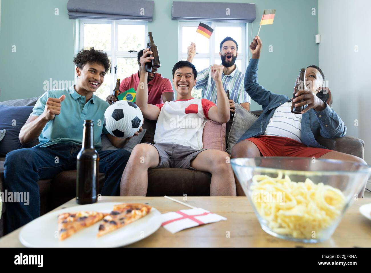 Amici maschili multirazziali urlano e sventolano bandiere mentre guardano la partita di calcio insieme a casa Foto Stock
