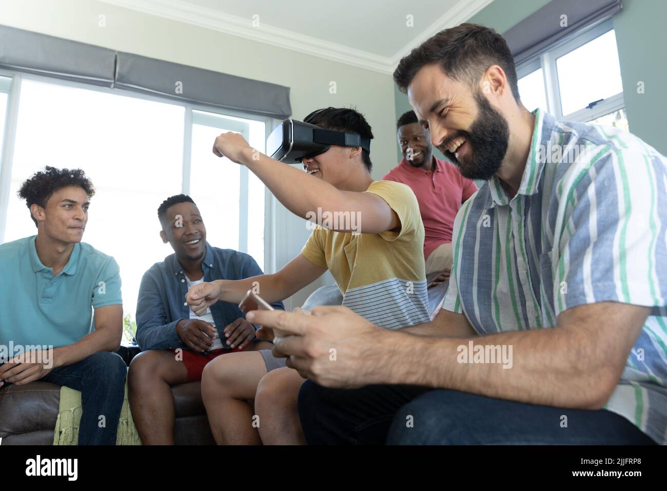 Amici multirazziali ridendo e guardando l'uomo che indossa un simulatore di realtà virtuale gesturing a casa Foto Stock