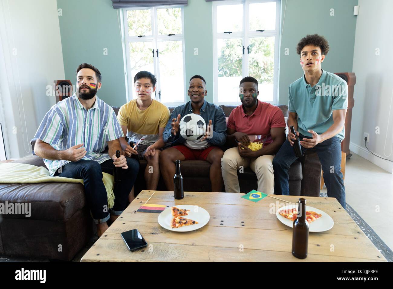 Curiosi amici maschi multirazziali con bottiglie di birra e pizza a tavola guardando partita di calcio a casa Foto Stock