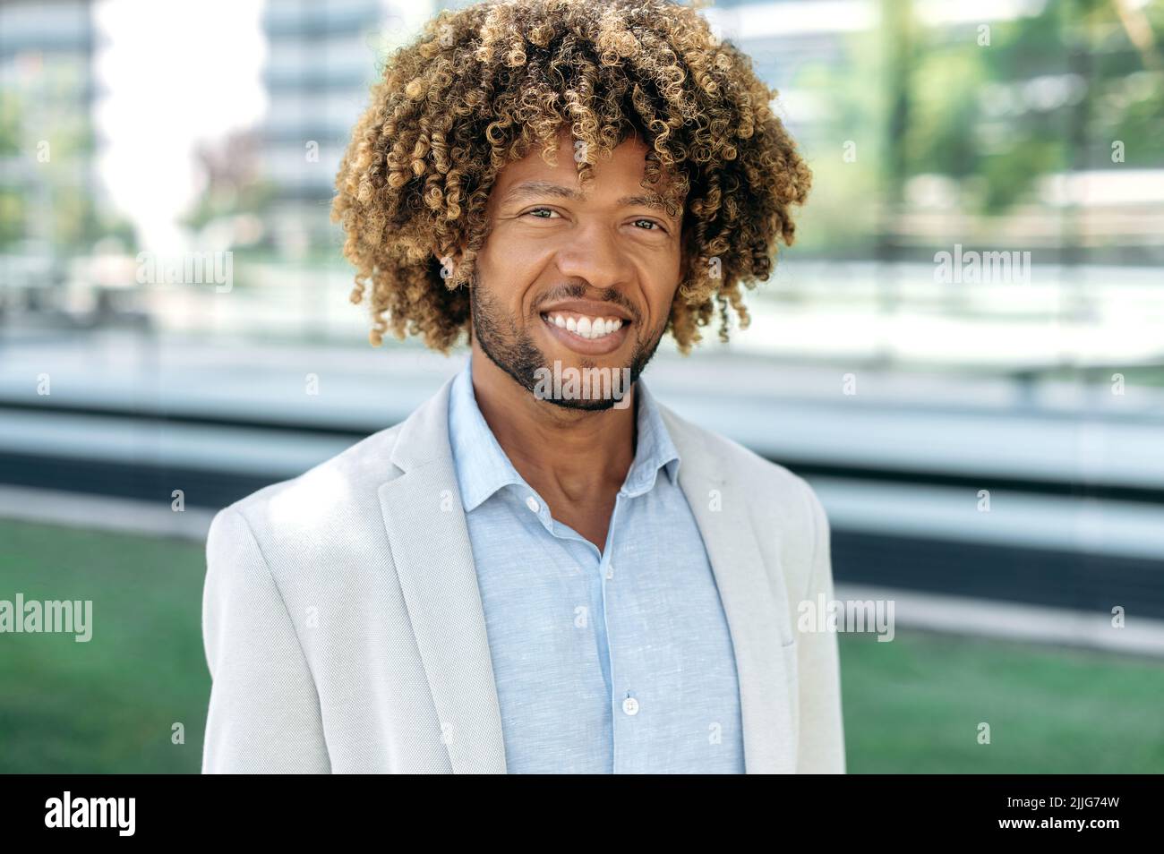 Primo piano di un bell'attraente afroamericano positivo o brasiliano uomo con capelli ricci, vestito in elegante abbigliamento formale, in piedi all'aperto, guardando la macchina fotografica, sorridente amichevole Foto Stock