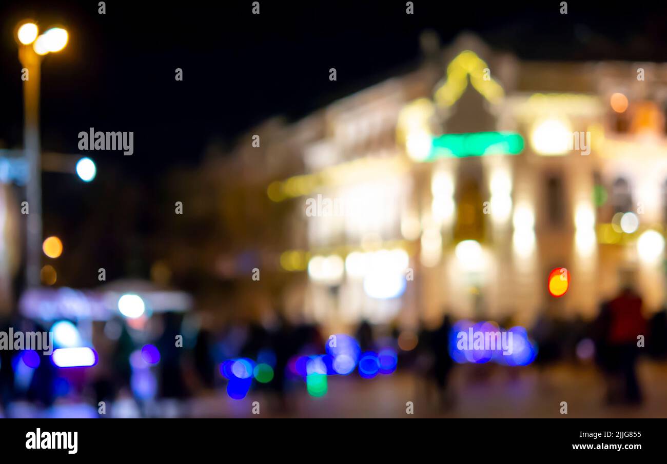 Sfondo sfocato. Silhouette nere sfocate delle persone, silhouette sfocata di un edificio architettonico illuminato da lanterne, retroilluminazione di notte. Piazza della città di sera. Molta gente sulla strada della città Foto Stock
