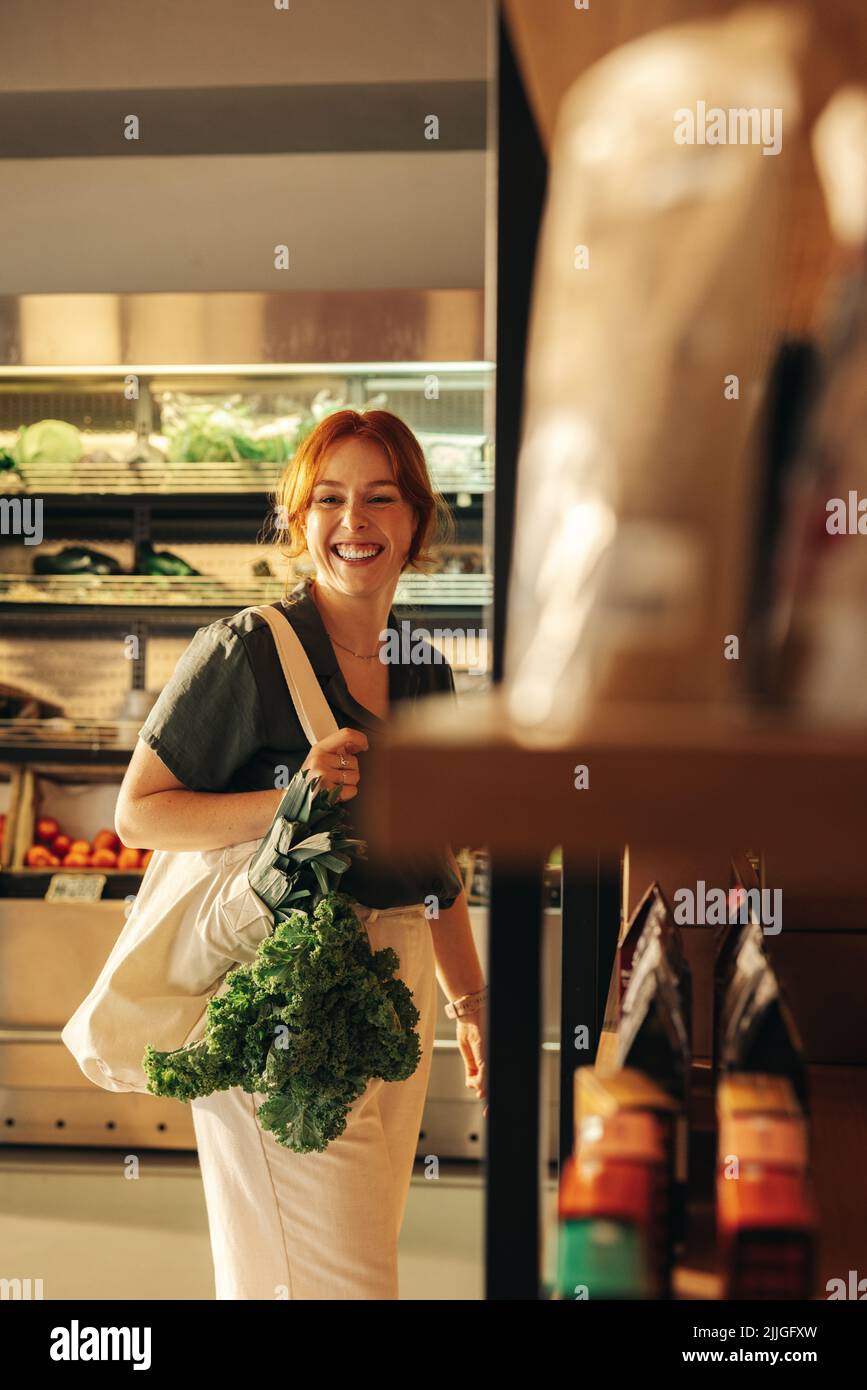 Cliente femminile che sorride felicemente mentre trasporta una borsa con verdure fresche biologiche in un negozio di alimentari. Allegro negozio di alimentari giovane donna in un tren Foto Stock