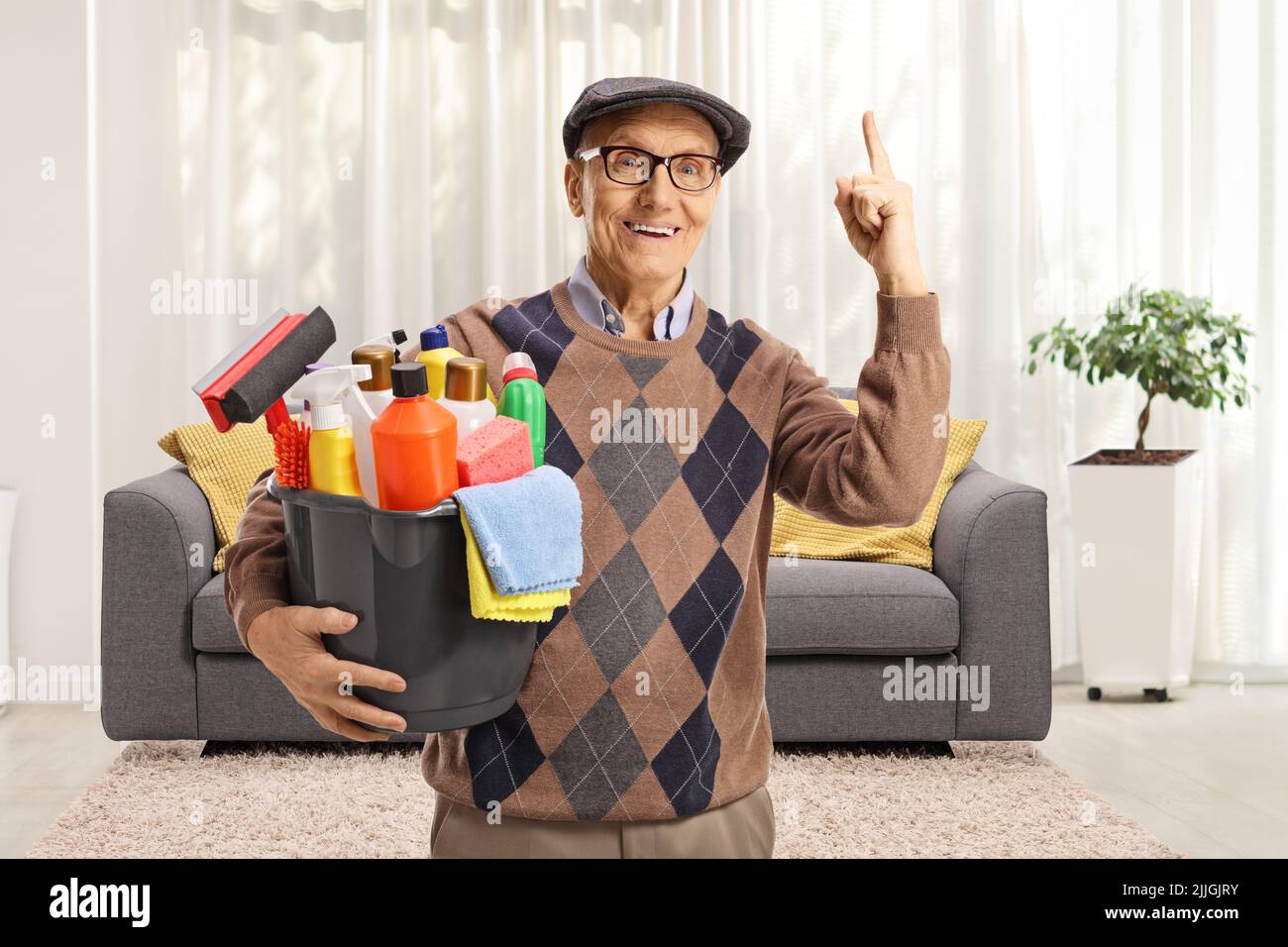 Uomo anziano con un secchio di forniture per la pulizia in piedi all'interno di un soggiorno e rivolto verso l'alto Foto Stock