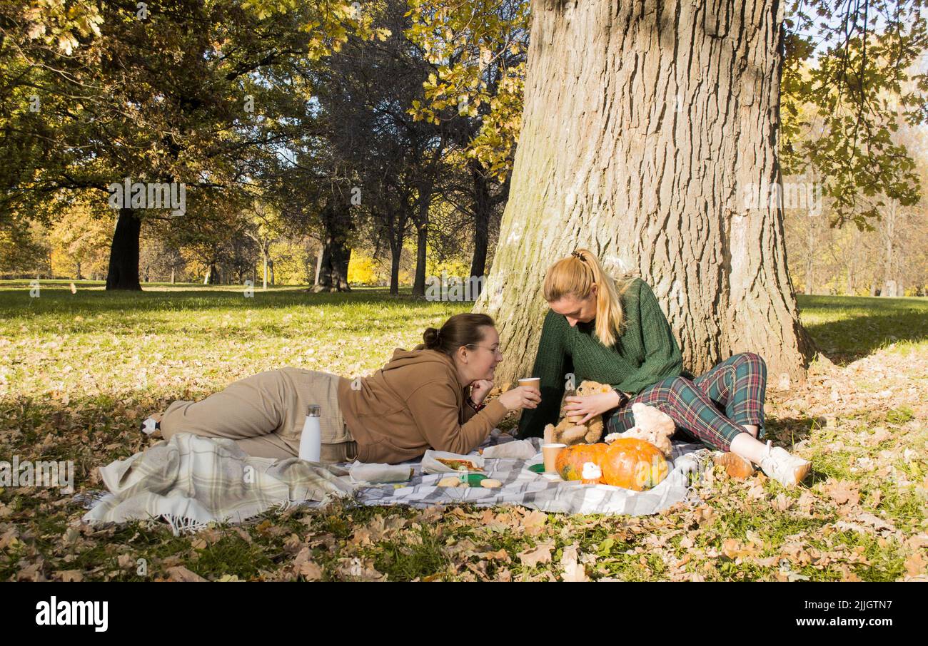 Due giovani donne felici in picnic in autunno parco seduto sotto albero su coperta, bere caffè, ridendo. Foto Stock