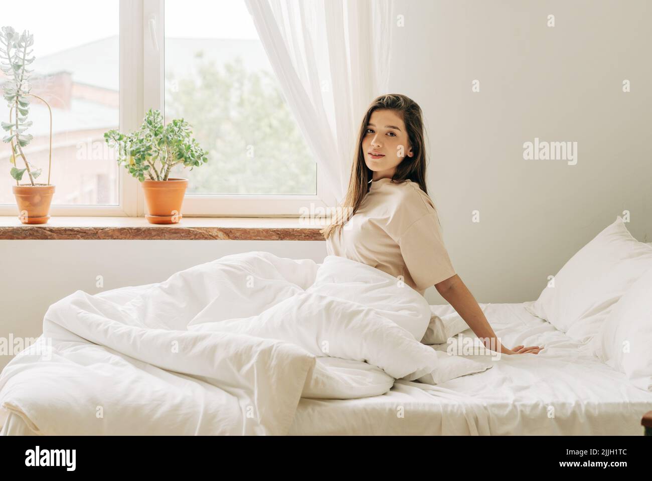 Giovane e attraente brunette si siede sul letto accanto alla finestra dopo il risveglio al mattino. Foto Stock