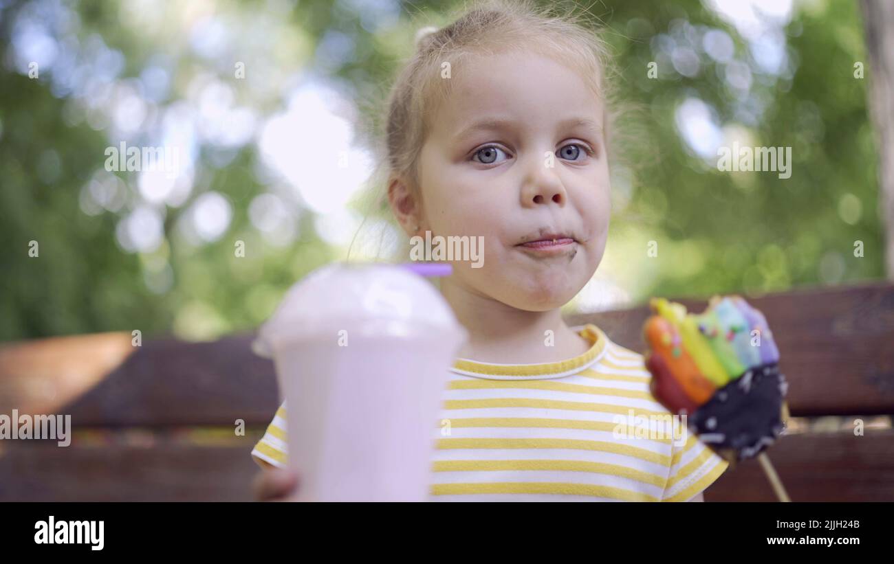 la ragazza della tartaruga mangia un pan di zenzero colorato e tiene un milkshake nella sua mano. Primo piano di ragazza carina bambino seduto sul panca del parco e mangiare biscotti con un Foto Stock