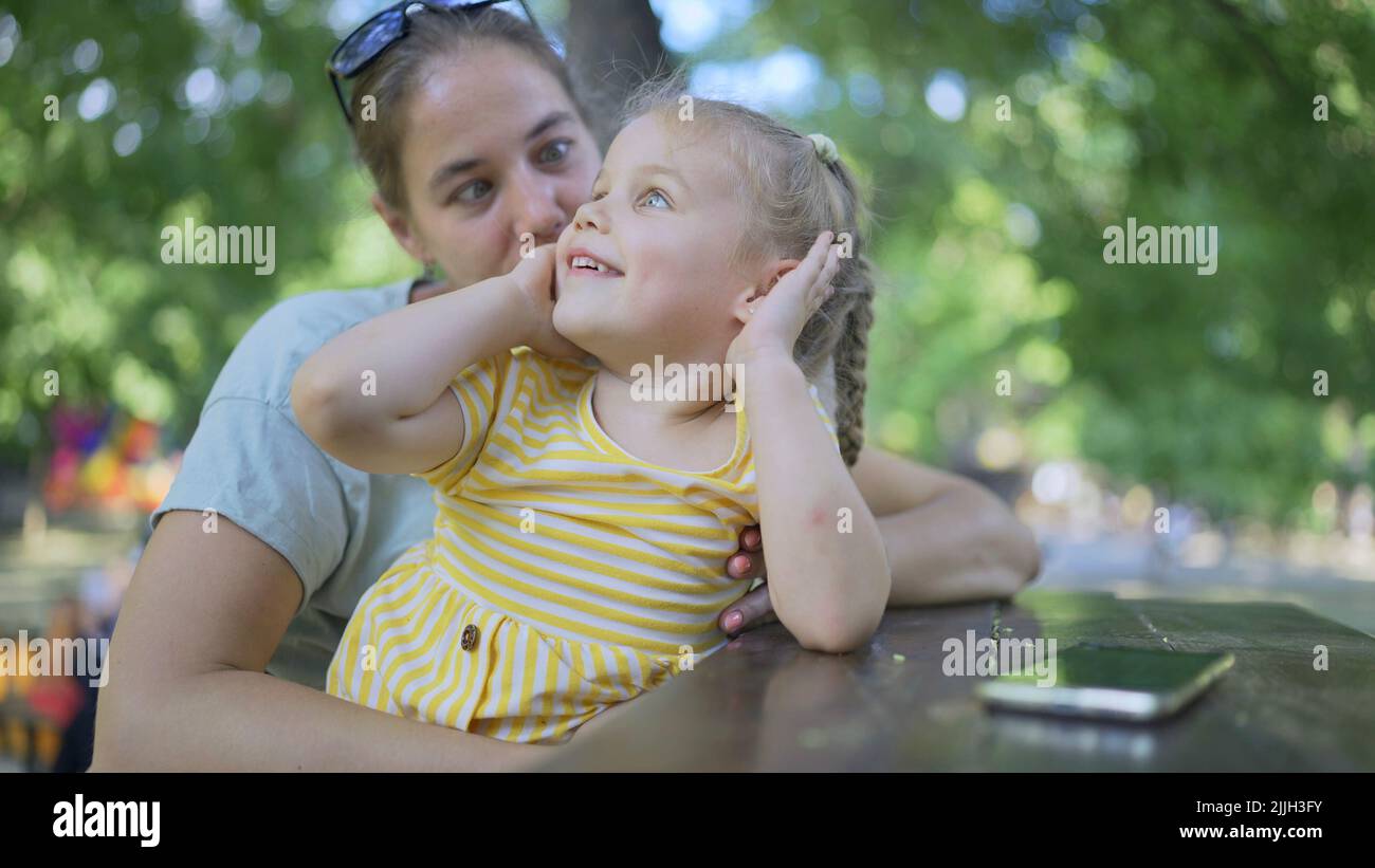 Primo piano di simpatica bambina gioca con la mamma mentre si siede nel parco. Primo piano di mamma e figlia mentre giocano seduti su una panchina del parco in una stree Foto Stock