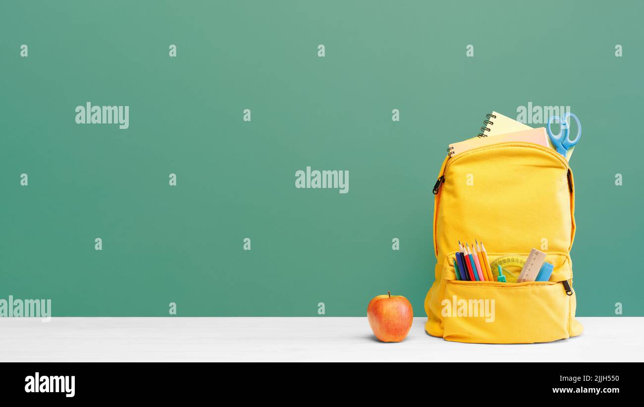 Zaino con diverse cancelleria colorata e mela sul tavolo e sfondo verde. Banner design back to School concept background. Foto Stock