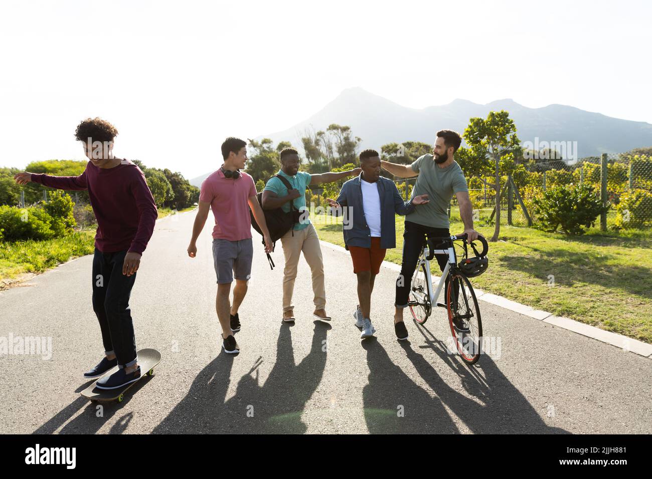 Amici maschi multirazziali skateboard, ciclismo e chiacchierare sulla strada contro alberi e cielo limpido Foto Stock