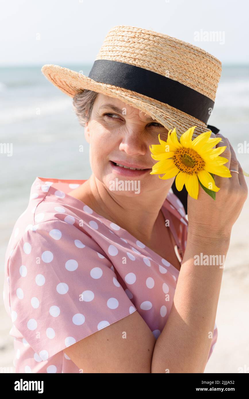 Felice sposata donna dai capelli grigi in un boater con un fiore giallo brillante, in un abito rosa con pallini bianchi che guardano qualcosa di interessante. Foto Stock
