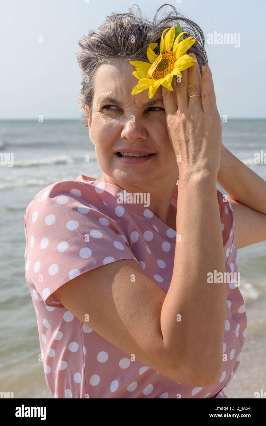 Donna sposata con capelli grigi in un abito rosa con puntini di polka bianco che tengono il fiore giallo nella sua mano rialzata guardando via. Concetto di beaty naturale. Foto Stock