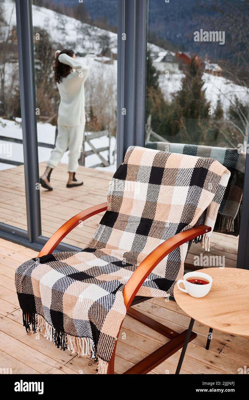Sedia e tavolo con una tazza di tè sulla terrazza della moderna casa di fienile in montagna d'inverno. Donna turistica si riflette in una finestra enorme. Foto Stock