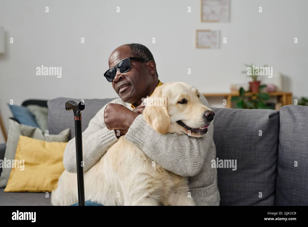 Uomo maggiore africano in occhiali con cattiva vista abbracciando il suo cane guida individuale mentre si siede sul divano a casa Foto Stock