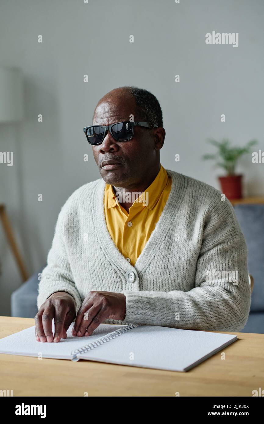 Cieco africano in vetri scuri seduti a tavola e libro di lettura in lingua braille Foto Stock