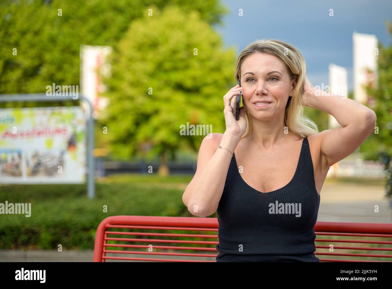 Posa naturale di una donna che chiacchiera sul suo smartphone all'aperto su una panca del parco e che corre la mano attraverso i suoi capelli Foto Stock