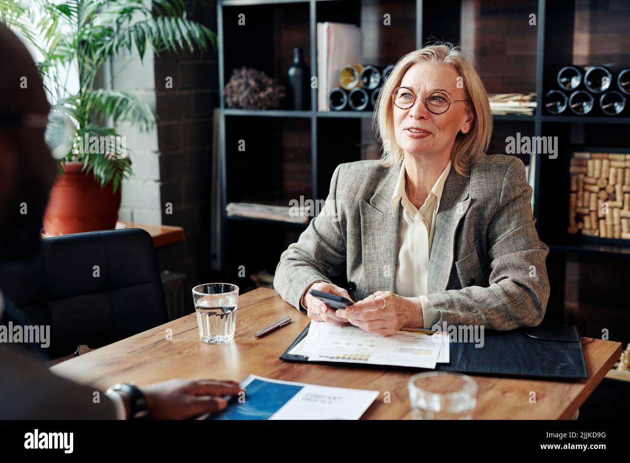 Donna d'affari matura in occhiali seduti al tavolo con documenti e utilizzando il suo telefono cellulare durante l'incontro con la collega al ristorante Foto Stock