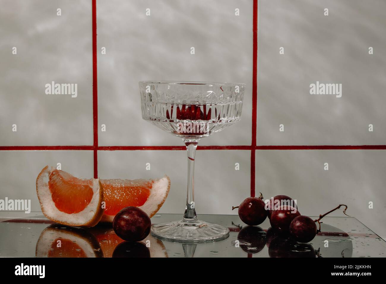 Vetro di cristallo per cocktail su una superficie di vetro. Frutta - uva, pompelmo accanto al bicchiere. Sfondo astratto. Foto Stock