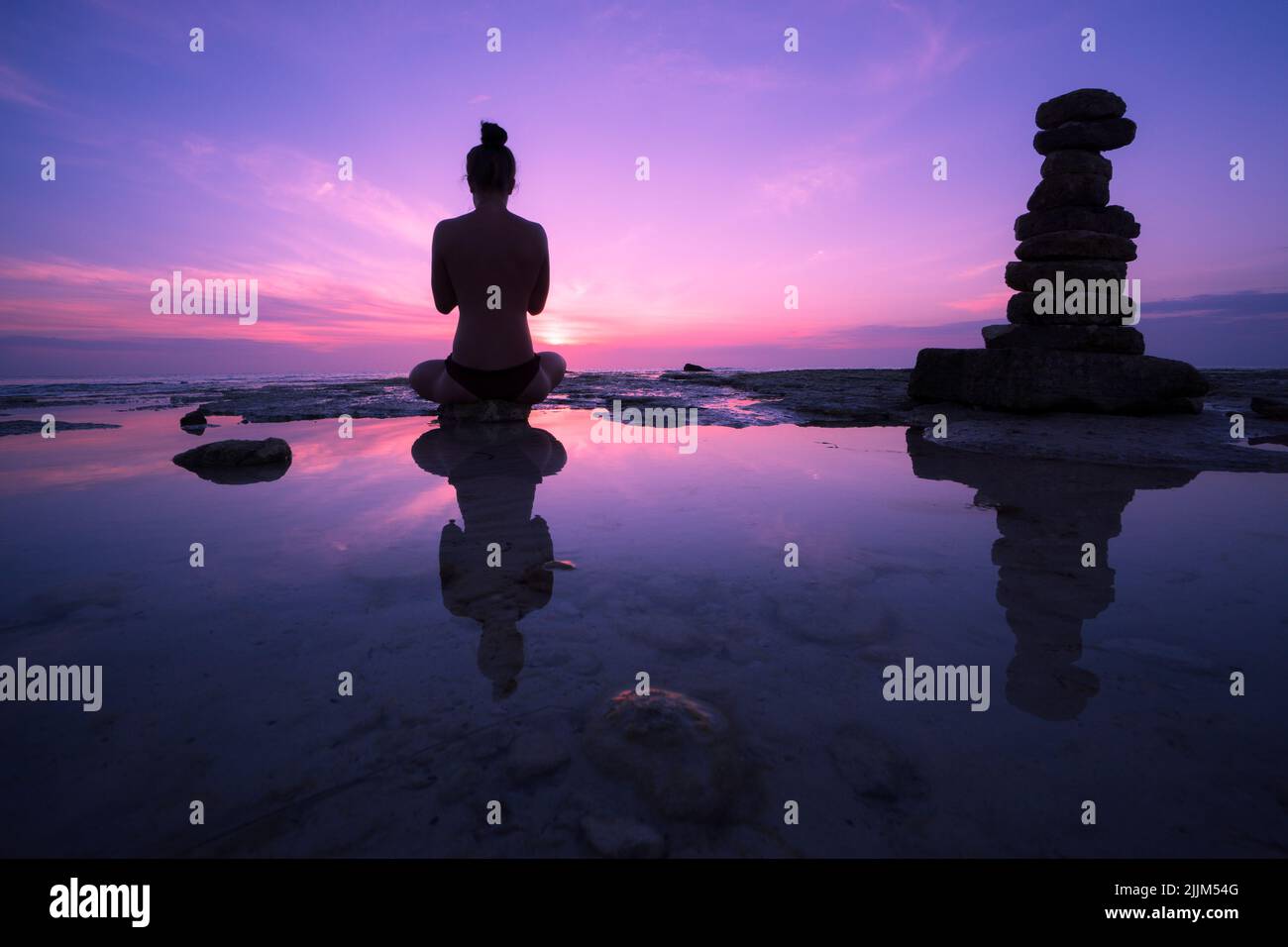 Una bella vista del tramonto in un colore viola e una donna meditating accanto all'acqua e alcune pietre Foto Stock