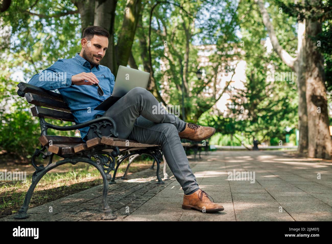 Un uomo d'affari giovane e sicuro che usa un notebook. I formali professionali maschili sono seduti sulla panca. Lavora al parco della città. Foto Stock