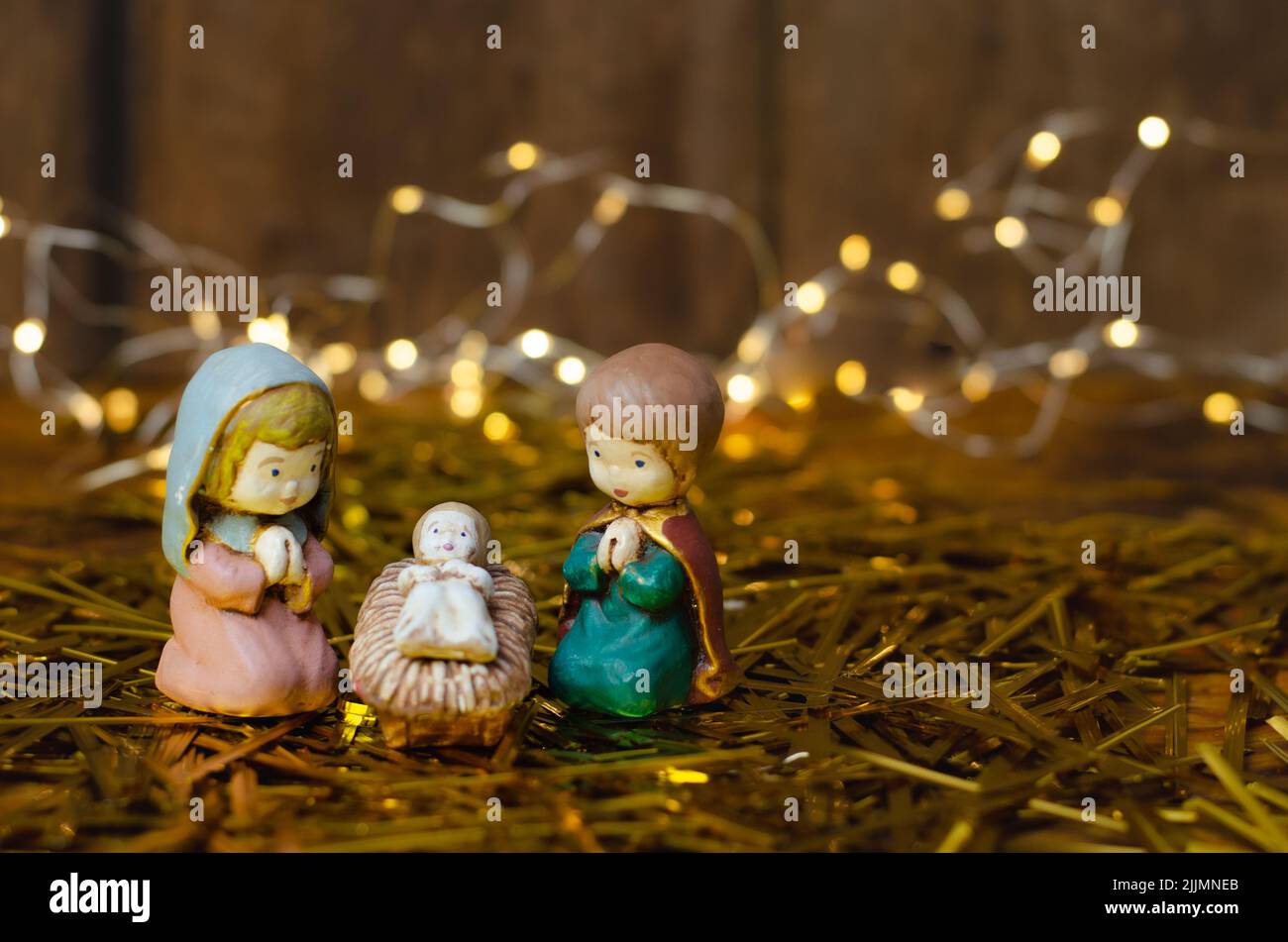 Un primo piano di mini figure colorate della scena Natività sul fieno con una serie di luci e una parete di legno marrone sullo sfondo sfocato Foto Stock