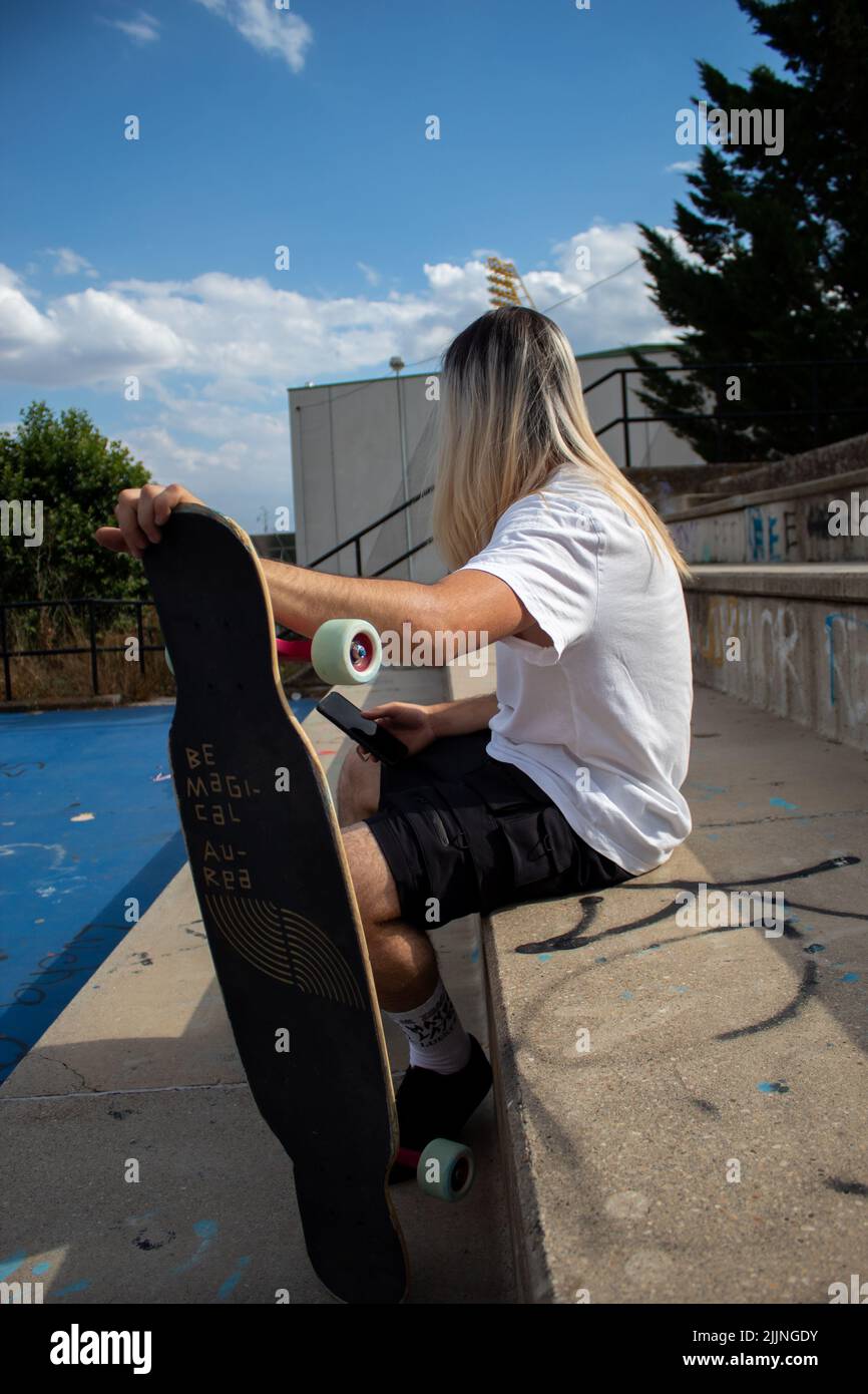 Una donna ispanica con capelli lunghi che tiene un longboard di legno seduto sui gradini e chiacchierando al telefono Foto Stock