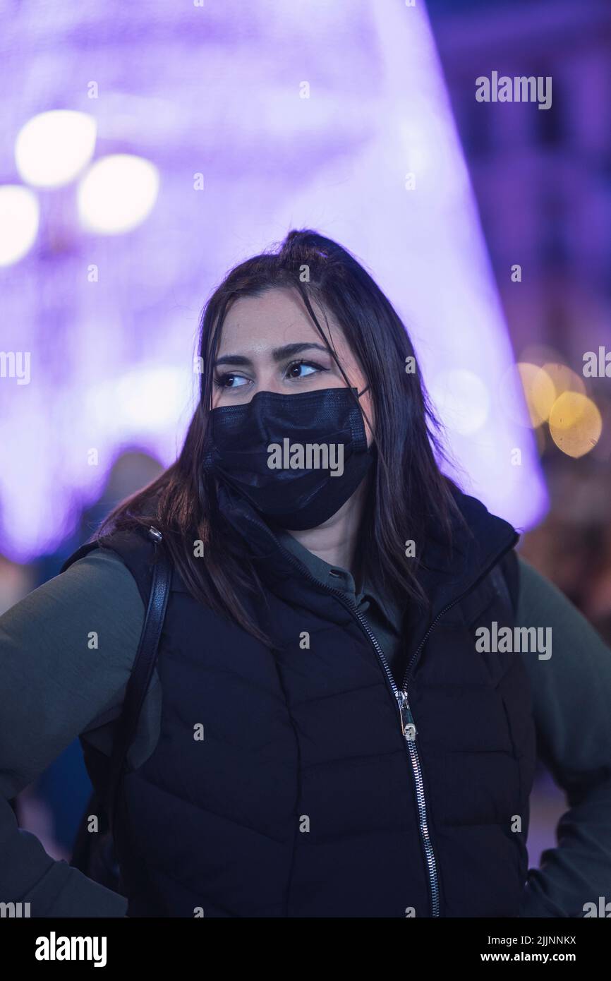 Una foto poco profonda di una femmina caucasica che indossa una giacca a maniche nere e una maschera nera con sfocate luci bokeh viola sullo sfondo Foto Stock