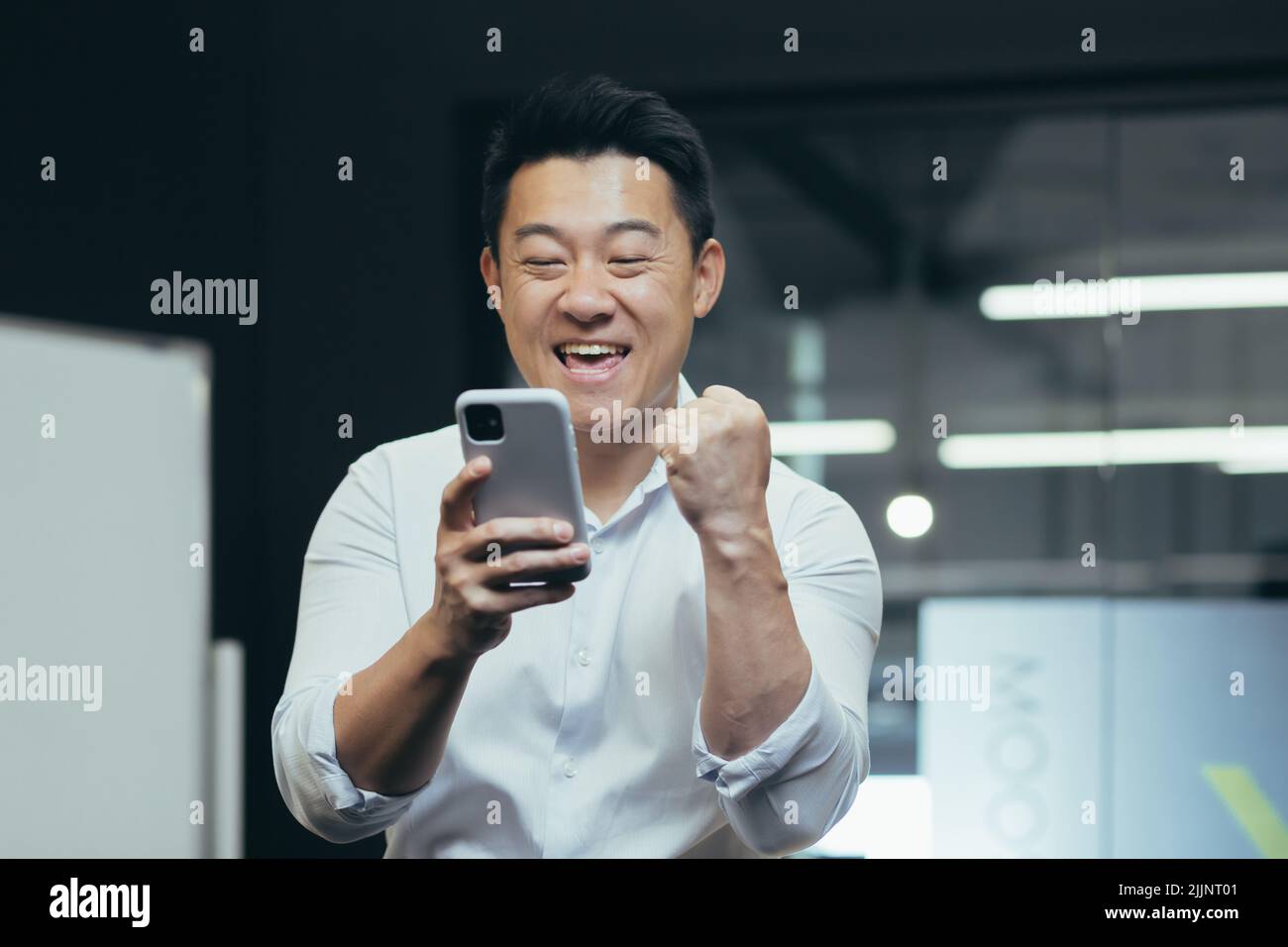 Una pausa al lavoro. Un giovane asiatico tiene un telefono nelle sue mani, gioca al telefono in ufficio al lavoro, si rallegra, mostra con la mano sì, si rallegra per la vittoria, ha vinto un premio, si riposa, non funziona. Foto Stock