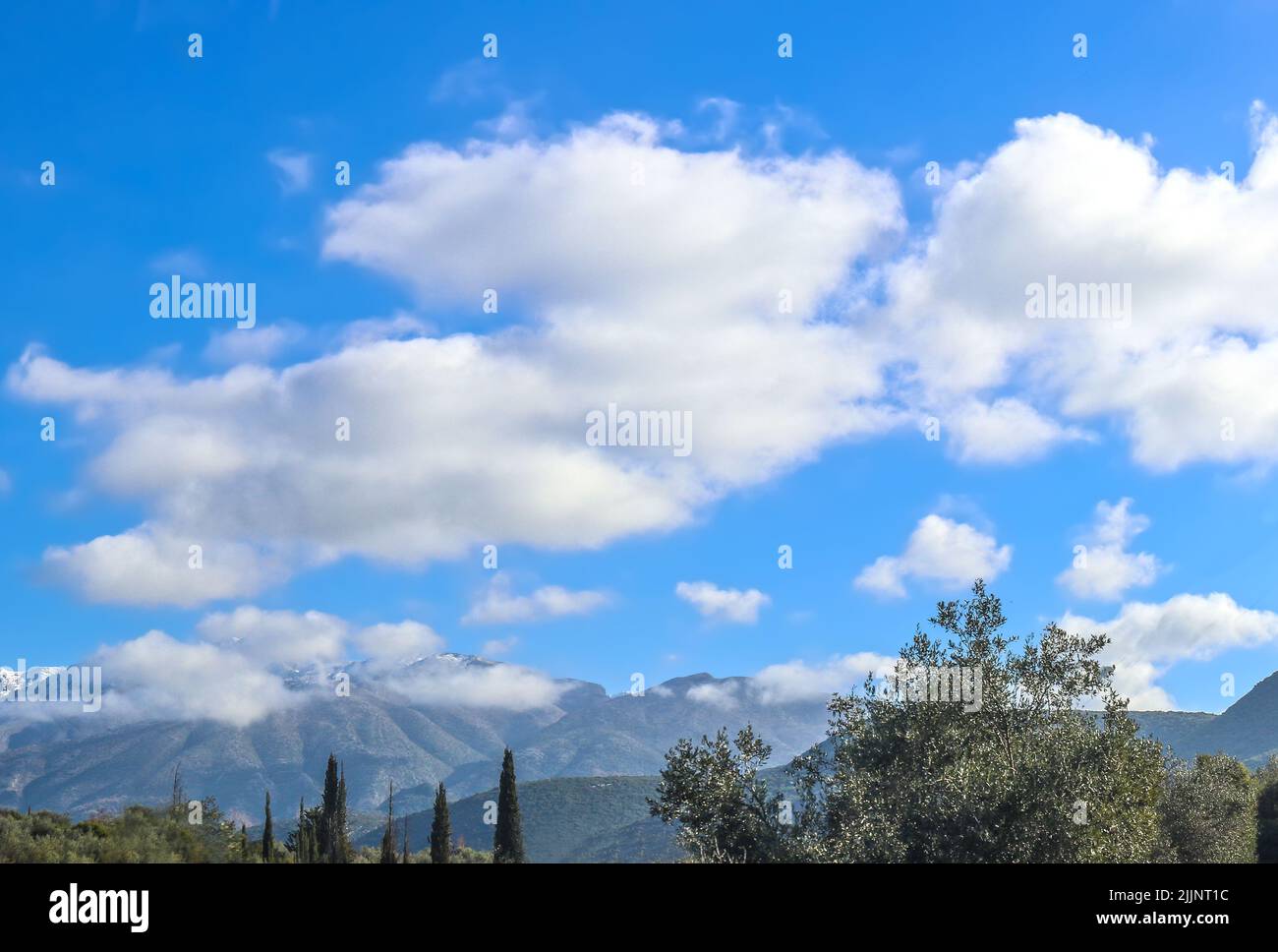 Monti greci del Peloponneso con neve e nubi cime coperte in lontananza sotto il cielo nuvoloso blu con olivi e cipressi in primo piano - s Foto Stock