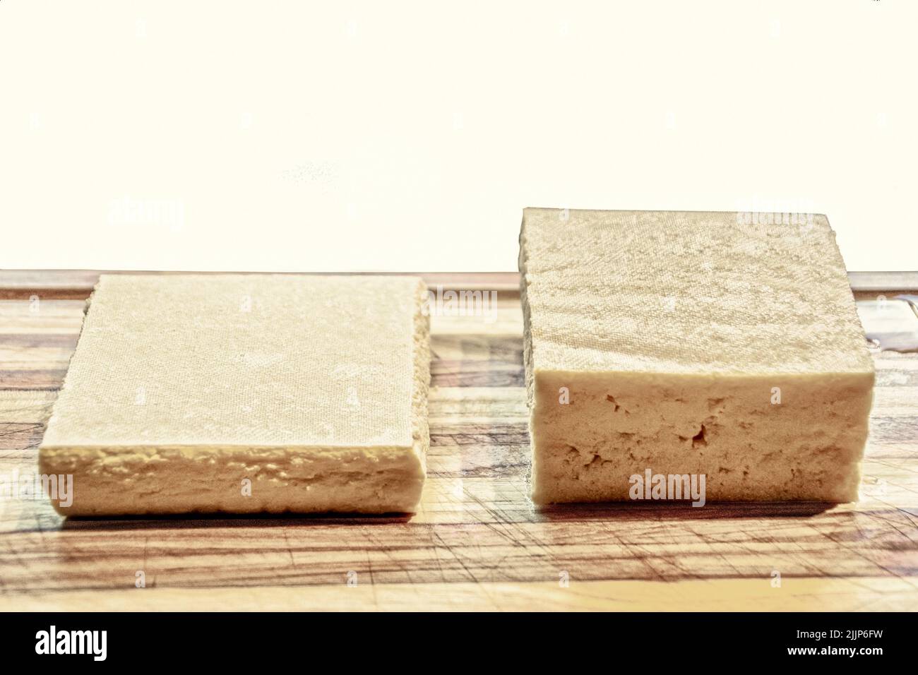 Due pezzi di tofu - uno fuori dalla confezione e l'altro premuto per rimuovere l'acqua - seduta su un tagliere con sfondo bianco - stanza per co Foto Stock