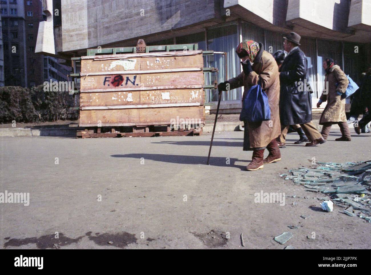 Bucarest, Romania, gennaio 1989. Persone che passano da vetri rotti e detriti in Piazza dell'Università, dall'Intercontinental Hotel, giorni dopo la rivoluzione anti-comunista del dicembre 1989. Una donna anziana sta coprendo il suo volto con un fazzoletto. Sul pannello di legno qualcuno ha scritto 'F.S.N.' (Fronte di salvezza nazionale - il partito che afferrò il potere dopo che Ceausescu fu eliminato), con il martello comunista e la falce in esso, accennando all'inclinazione del partito. Foto Stock