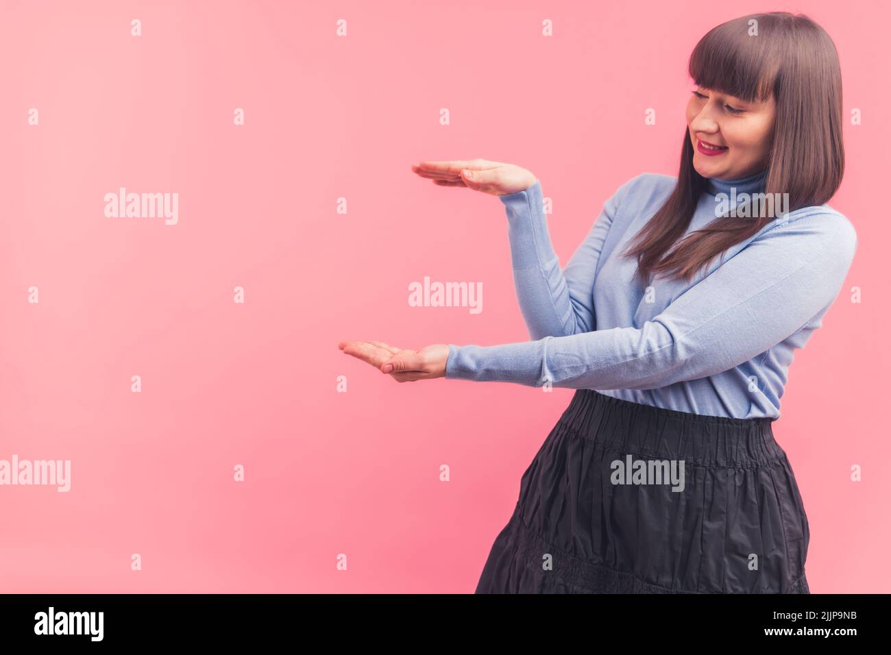 Giovane donna bruna caucasica con i bangs che indossano un dolcevita blu che incornicia con le mani e guarda a sinistra. Foto studio con sfondo rosa. . Foto di alta qualità Foto Stock