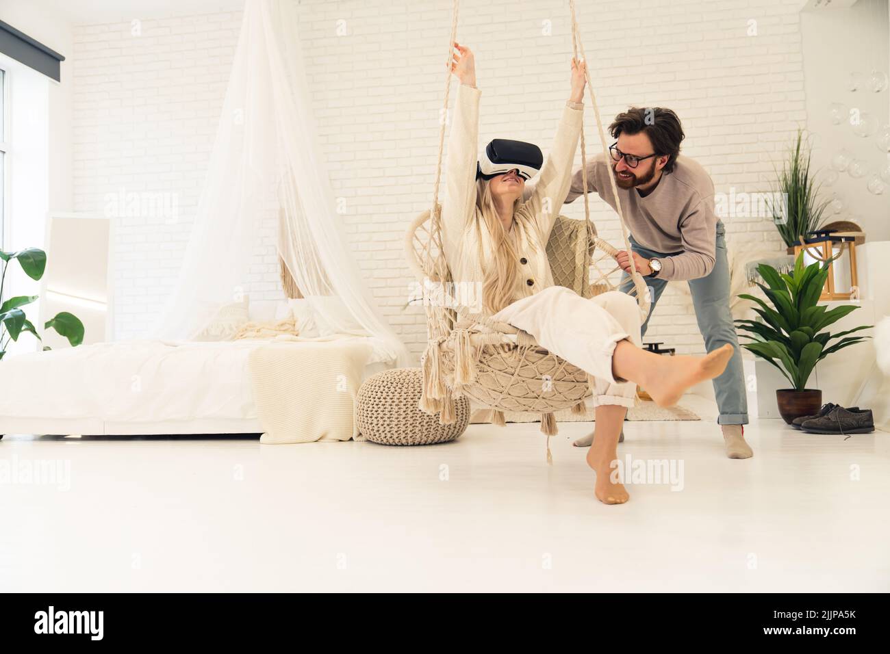 Giovane coppia bianca in camera da letto stile boho. Donna bionda in occhiali VR su sedia sospesa tenuta da uomo con barba e occhiali scuri. Scatto al coperto. Foto di alta qualità Foto Stock