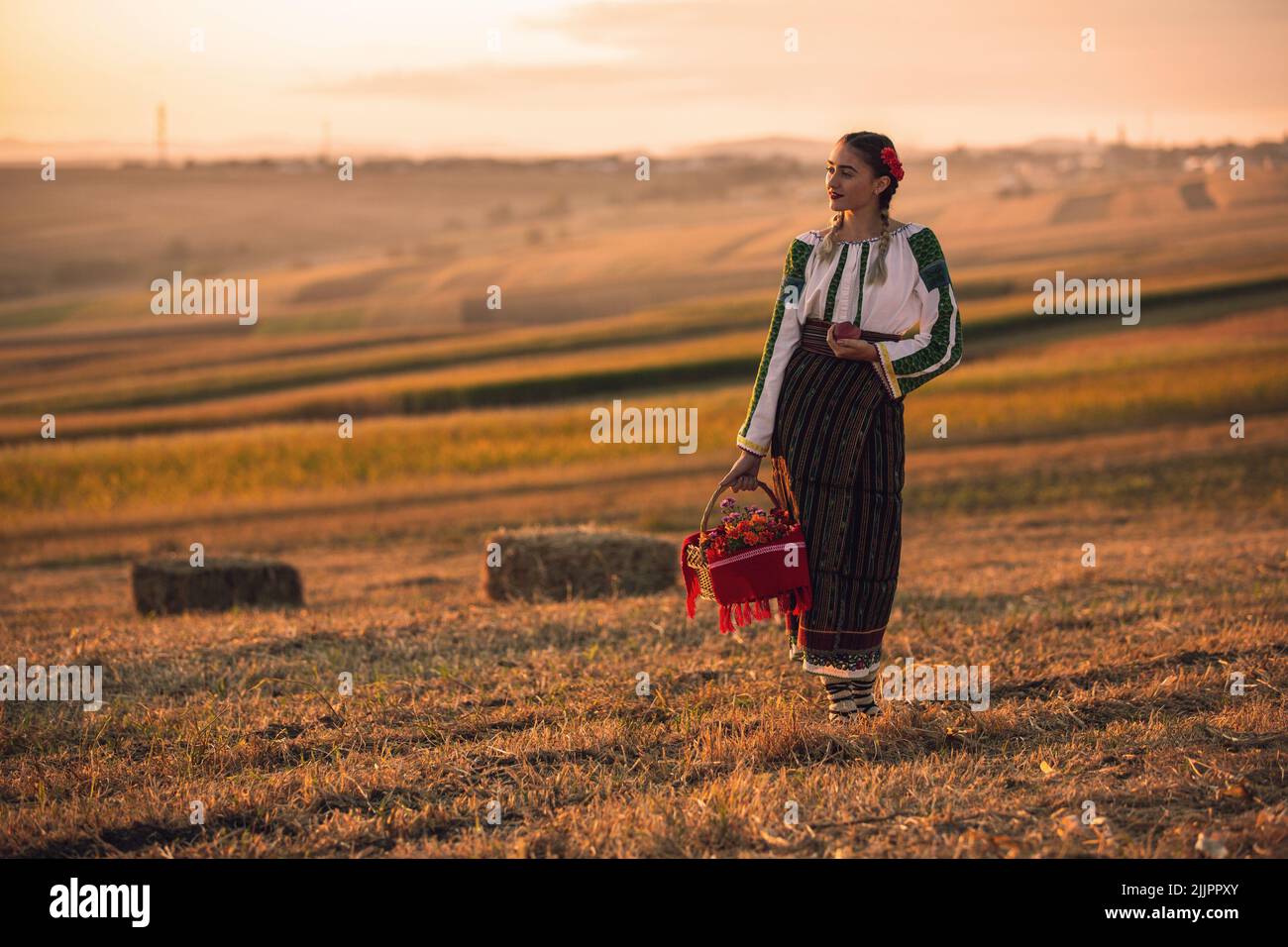 Una giovane donna rumena in costume tradizionale e cesto che si posa sul campo al tramonto panoramico Foto Stock