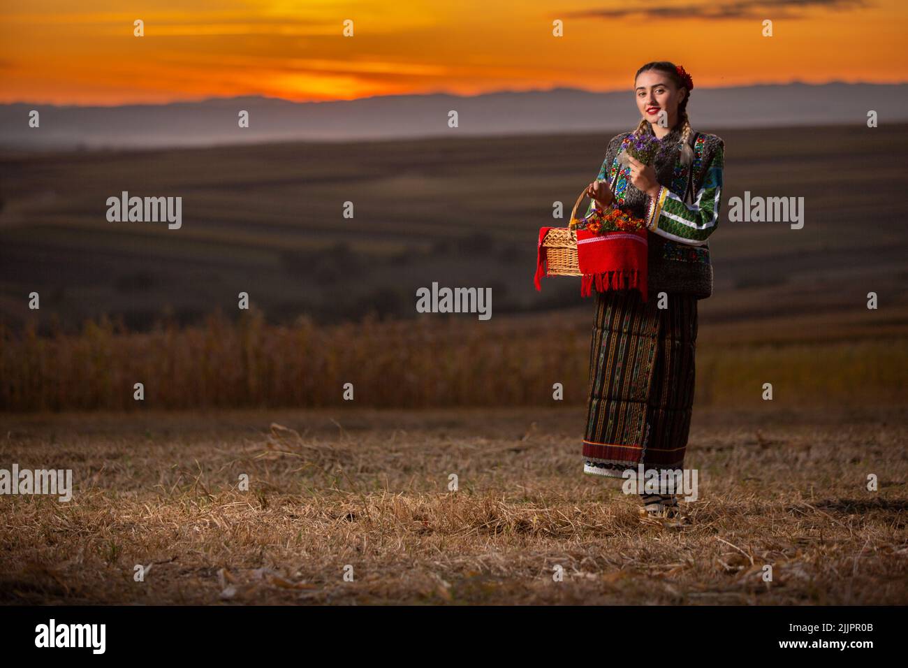 Una giovane donna rumena in costume tradizionale e cesto che si posa all'aperto al tramonto panoramico Foto Stock