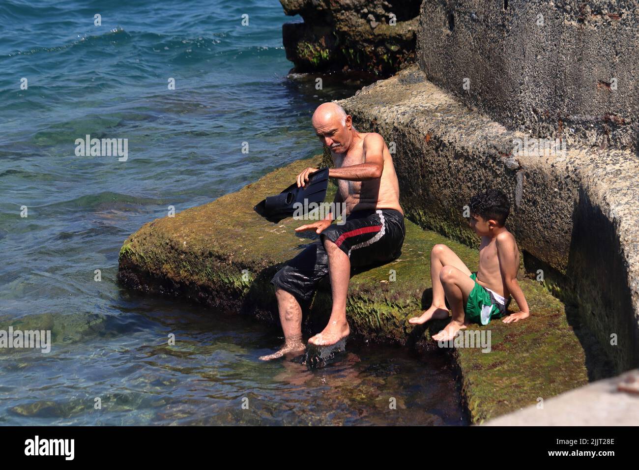 Il vecchio e il ragazzo si siedono sulle rocce dopo lo snorkeling in mare. Forse nonno e nipote. Nonno ha appena rimosso il flipper. Ragazzo lo ammira. Foto Stock