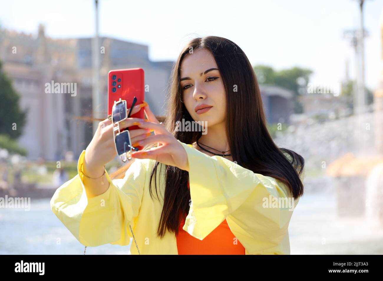 Ragazza attraente con capelli lunghi e trucco perfetto prendendo selfie su smartphone fotocamera in piedi su una strada. Bellezza femminile, turista in città d'estate Foto Stock