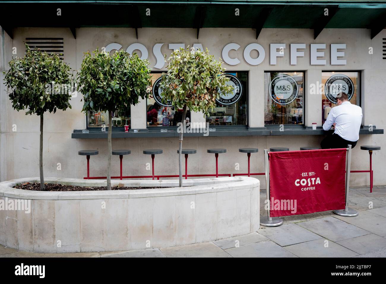 Accanto a tre arbusti che si appassiscono per la mancanza di acqua, un uomo siede su uno sgabello in un bar da asporto Costa Coffee nella City di Londra, il quartiere finanziario della capitale, il 27th luglio 2022, a Londra, Inghilterra. Costa Coffee è stata fondata a Londra nel 1971 da Sergio Costa - acquisita da Whitbread nel 1995 poi venduta nel 2019 alla Coca-Cola Company in un accordo del valore di £ 3,9bn. La sua attività nel Regno Unito ha 2.121 ristoranti nel Regno Unito, oltre 6.000 distributori automatici Costa Express e altri 1.280 punti vendita all'estero. Foto Stock