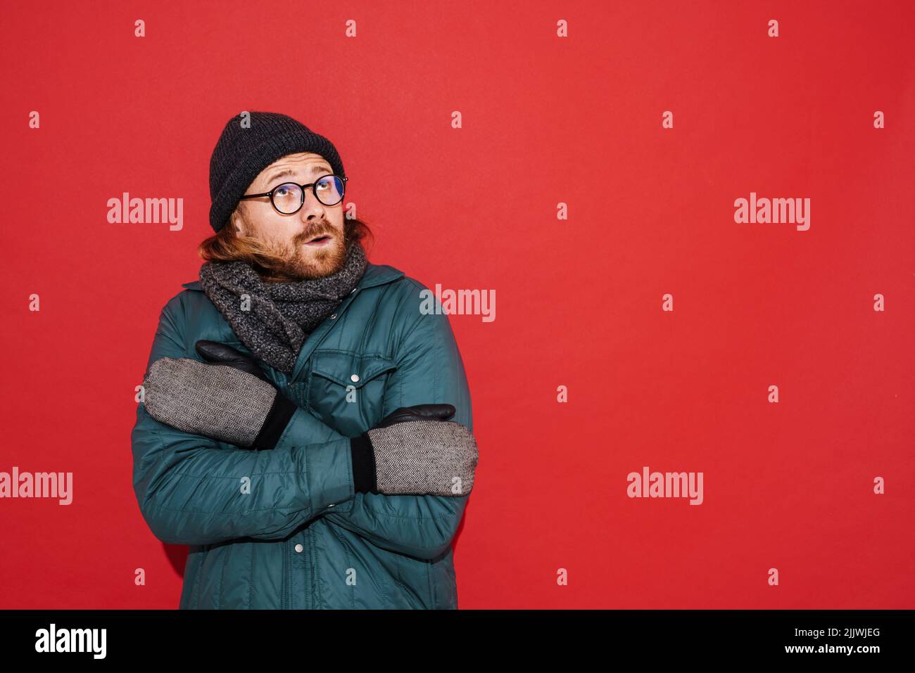 Uomo zenzero che indossa cappello e giacca tremante e sensazione di freddo isolato su sfondo rosso Foto Stock