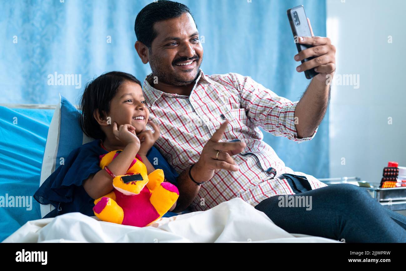 Felice padre sorridente con la figlia ammalata ricuperata che fa videochiamata sul telefono cellulare al reparto dell'ospedale - concetto di tecnologia, rapporto e salute Foto Stock