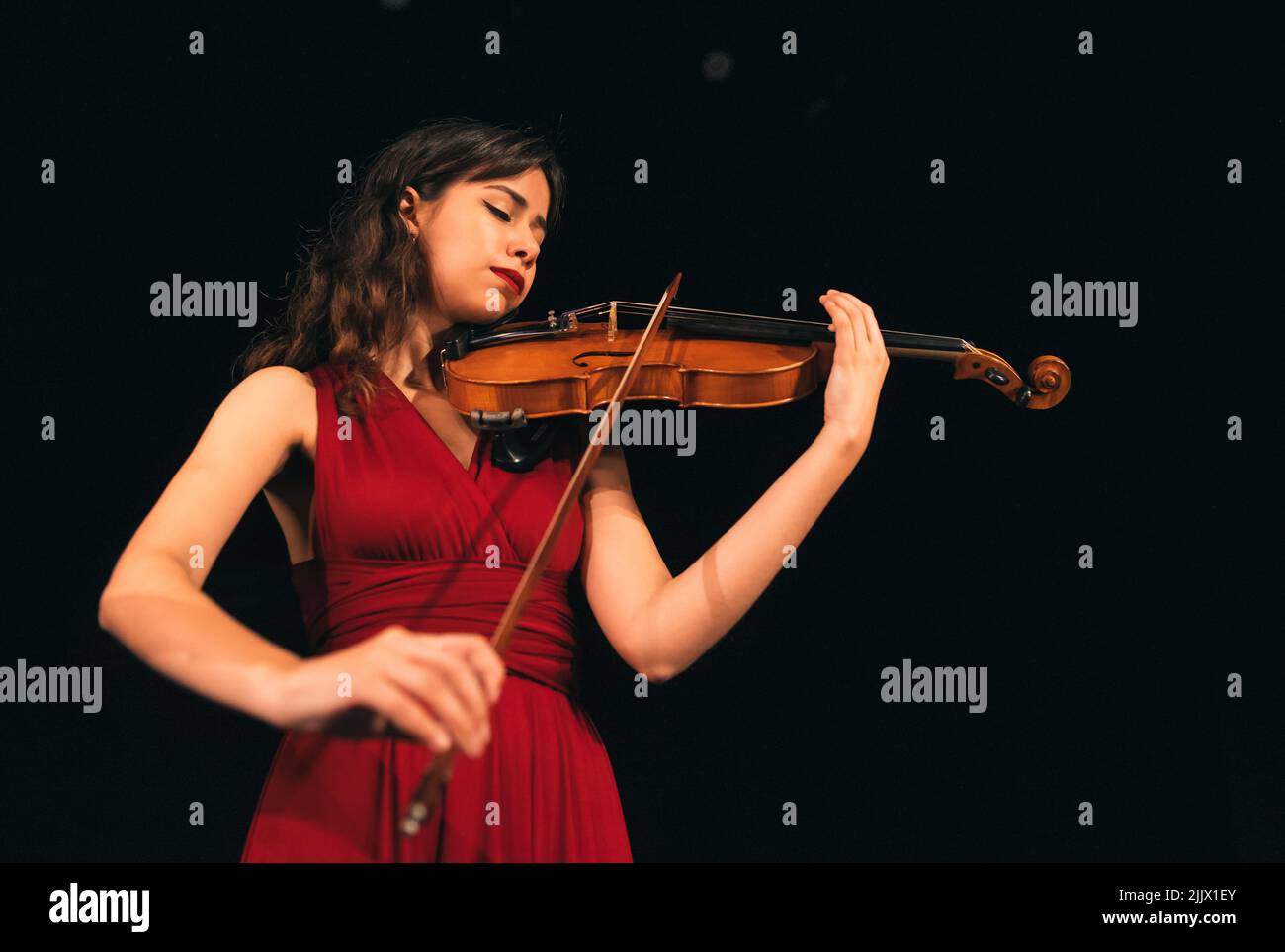 Da sotto Donna in abito rosso suonando violino con occhi chiusi durante il concerto sul palco in teatro scuro Foto Stock