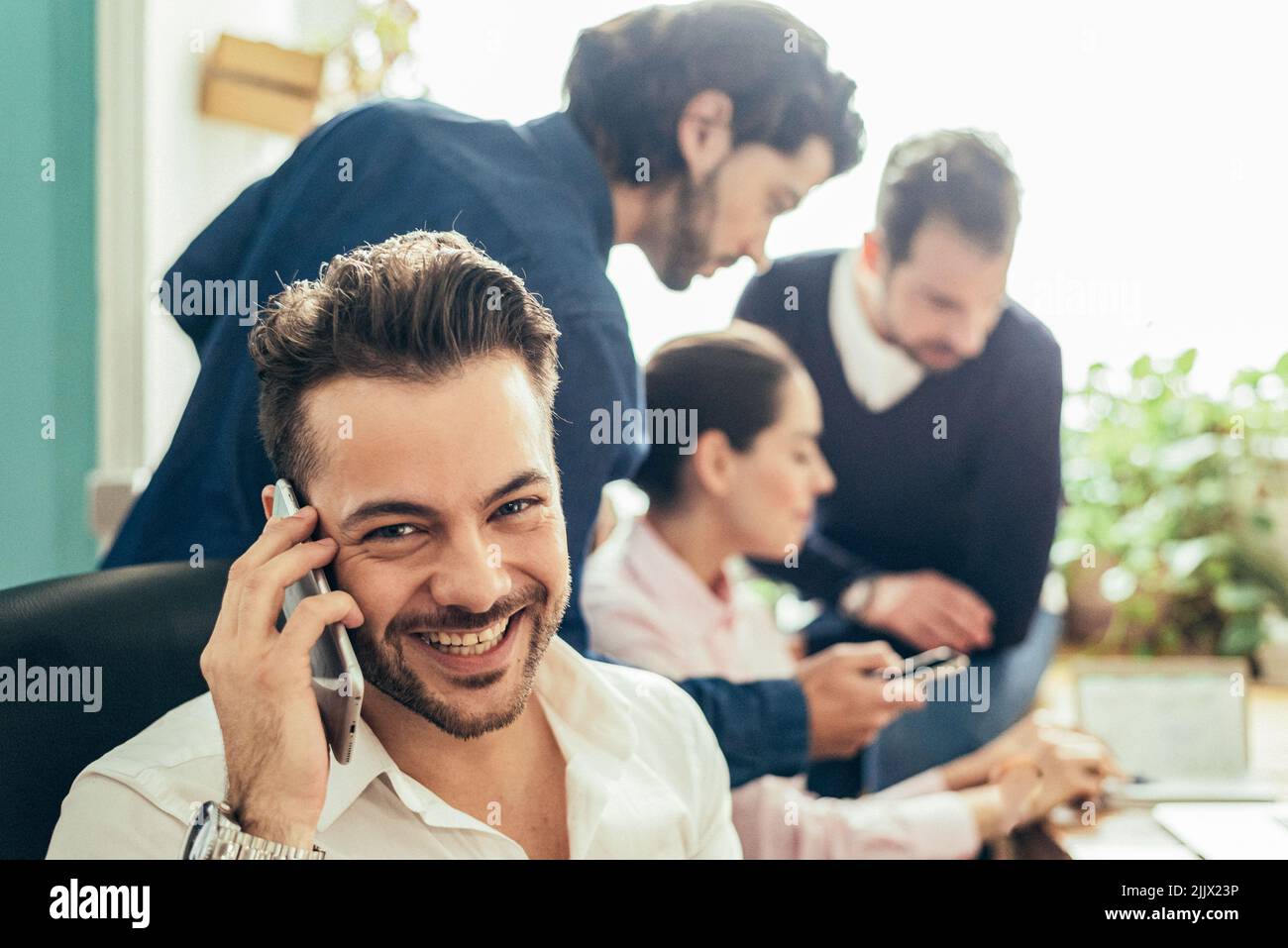 buon lavoratore di sesso maschile in abbigliamento formale che ha una conversazione telefonica vicino a un gruppo di colleghi mentre lavora insieme in un moderno ufficio leggero Foto Stock