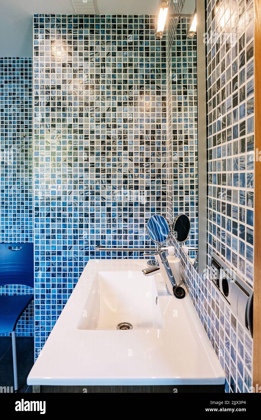 Lavandino in ceramica bianca con rubinetto cromato e specchio cosmetico in bagno contemporaneo con pareti piastrellate in mosaico blu Foto Stock