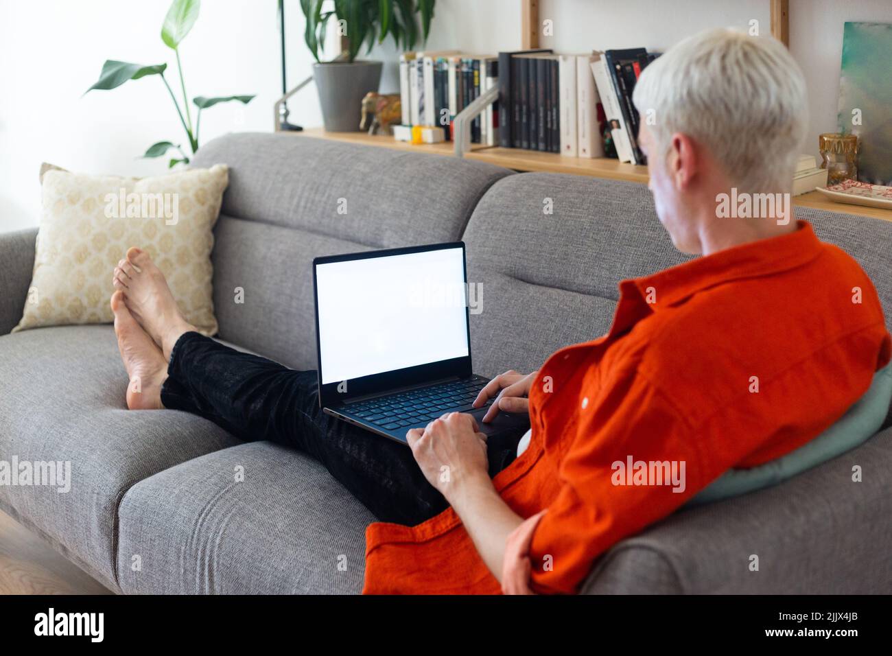Corpo completo di uomo transgender utilizzando un computer portatile seduto sul divano in soggiorno Foto Stock