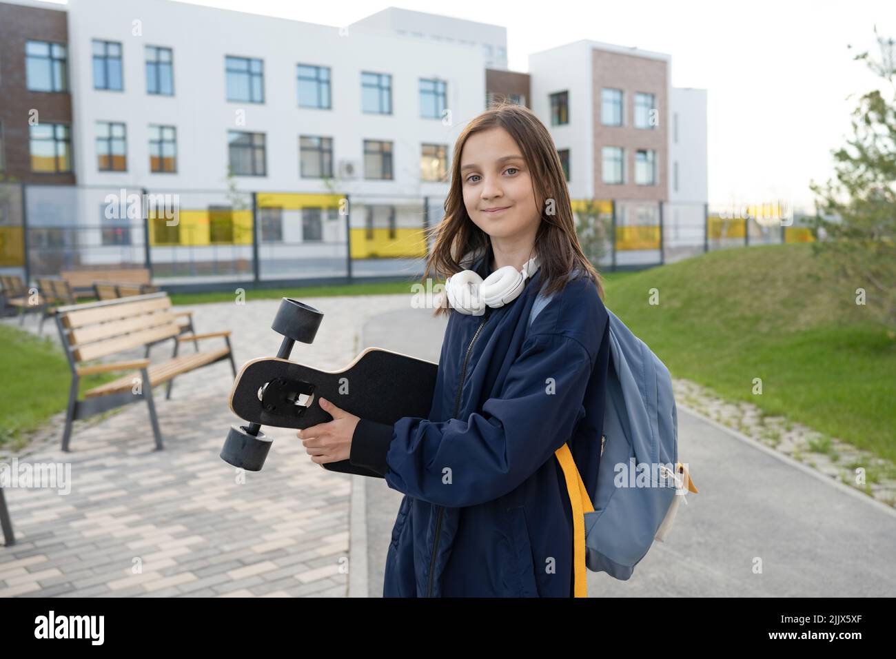 Ragazza premedetta in abbigliamento casual con longboard passeggiando sulla passerella vicino a scuola il giorno d'estate in città guardando la macchina fotografica Foto Stock