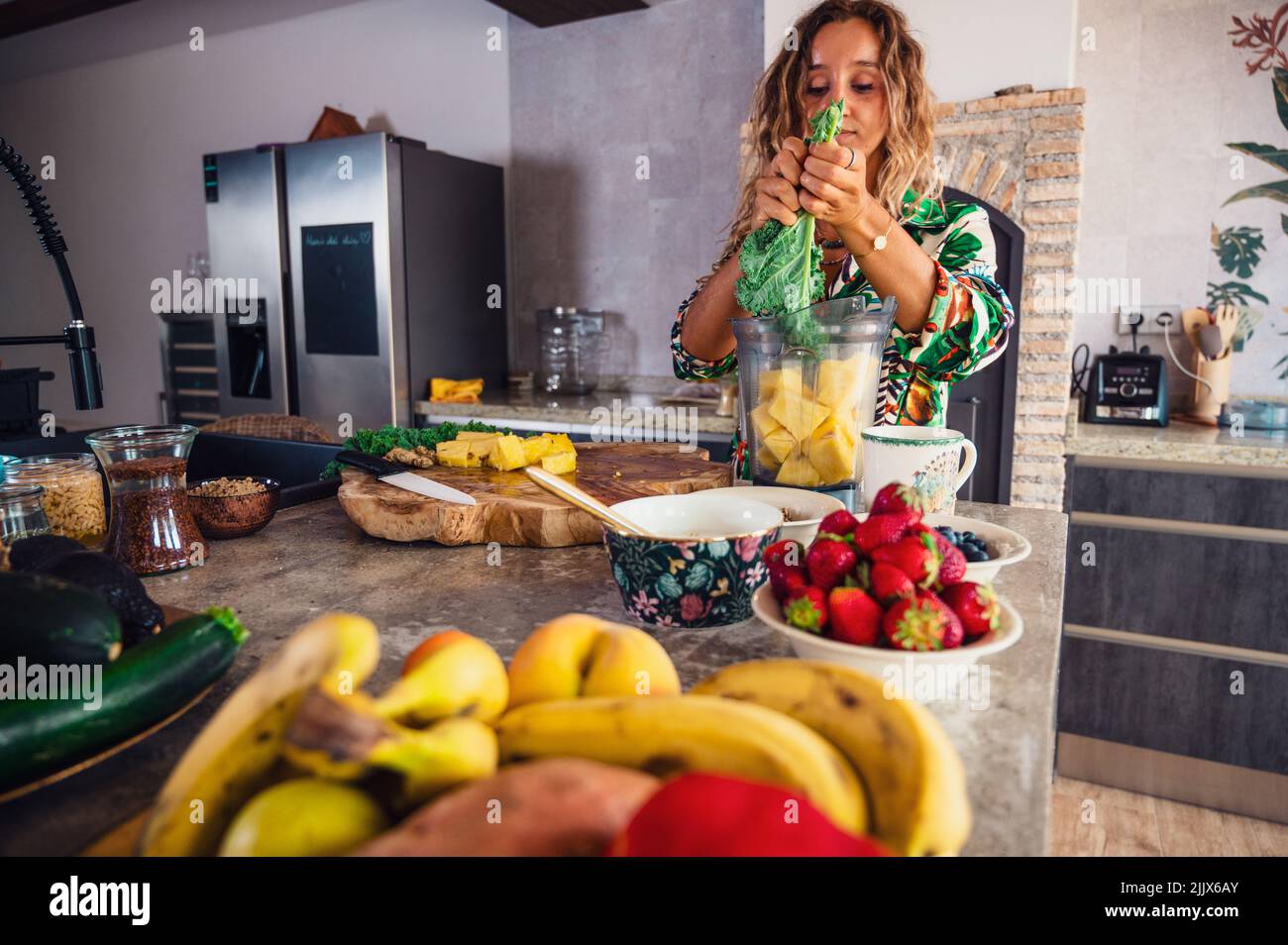 Donna che mette il kale verde nel recipiente del frullatore con ananas tritato mentre prepara il frullato al tavolo con frutta e bacche varie Foto Stock